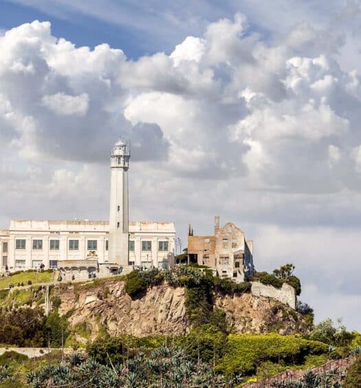 A Ilha de Alcatraz com seu prédio principal da prisão e o farol sob um céu parcialmente nublado, cercado por vegetação. - Tour por São Francisco + Alcatraz