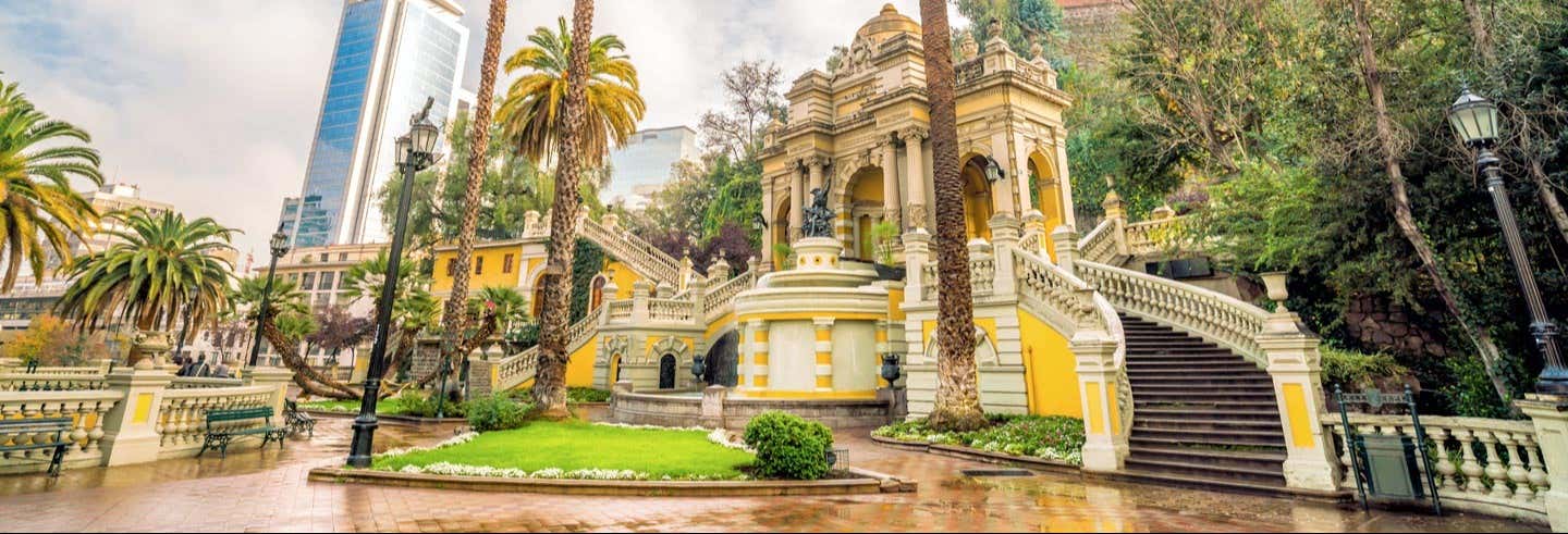 Edifício de pedra ornamentado em amarelo e branco com escadas, palmeiras e jardins em um parque urbano. A foto é capa do post do Tour por Santiago + Teleférico del cerro San Cristóbal.