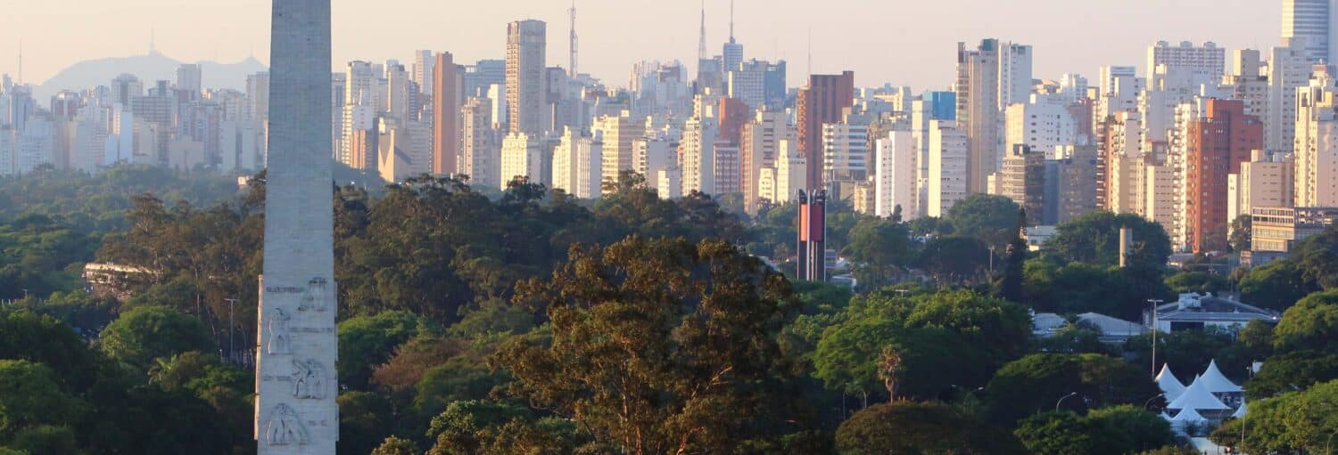 Prédios altos atrás de uma densa área de árvores com um grande monumento de pedra em primeiro plano ao pôr do sol. A imagem ilustra o post sobre Tour completo por São Paulo.