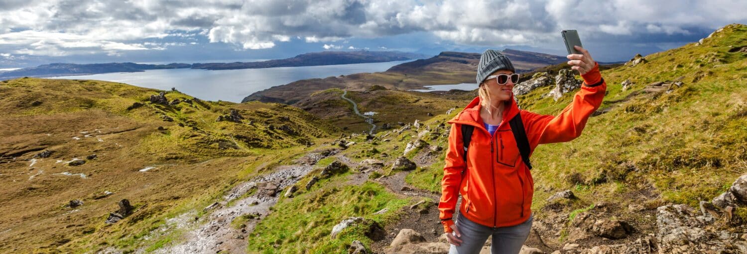 Uma pessoa com uma jaqueta laranja tira uma selfie em uma colina rochosa com água e montanhas ao fundo. A foto é capa do post do Tour de 3 dias a Skye e Highlands.