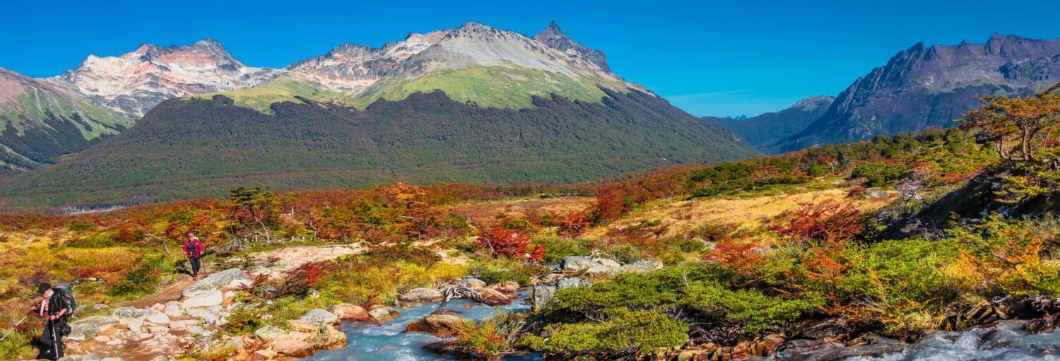 Dois caminhantes percorrem uma trilha rochosa ao lado de um riacho com montanhas e folhagens coloridas ao fundo, sob um céu azul. Imagem para ilustrar post sobre a Terra do Fogo + Trem do Fim do Mundo e Canal Beagle.