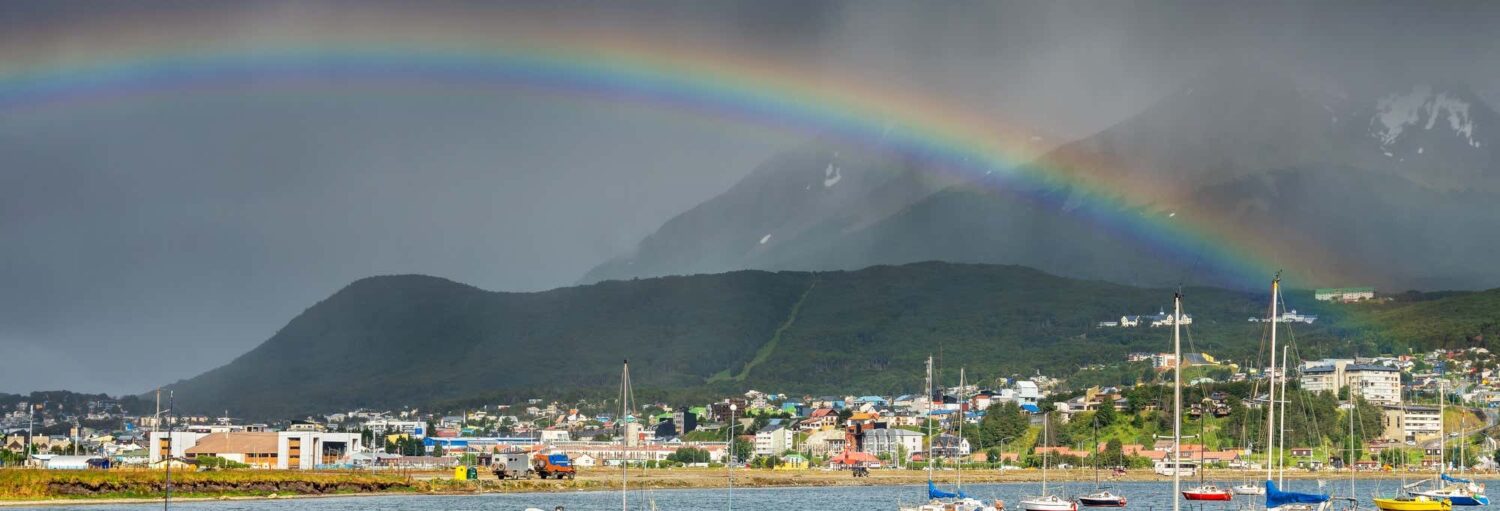 Um arco-íris se estende sobre uma cidade costeira com veleiros em primeiro plano e montanhas ao fundo. Imagem para ilustrar post sobre tour por Ushuaia.