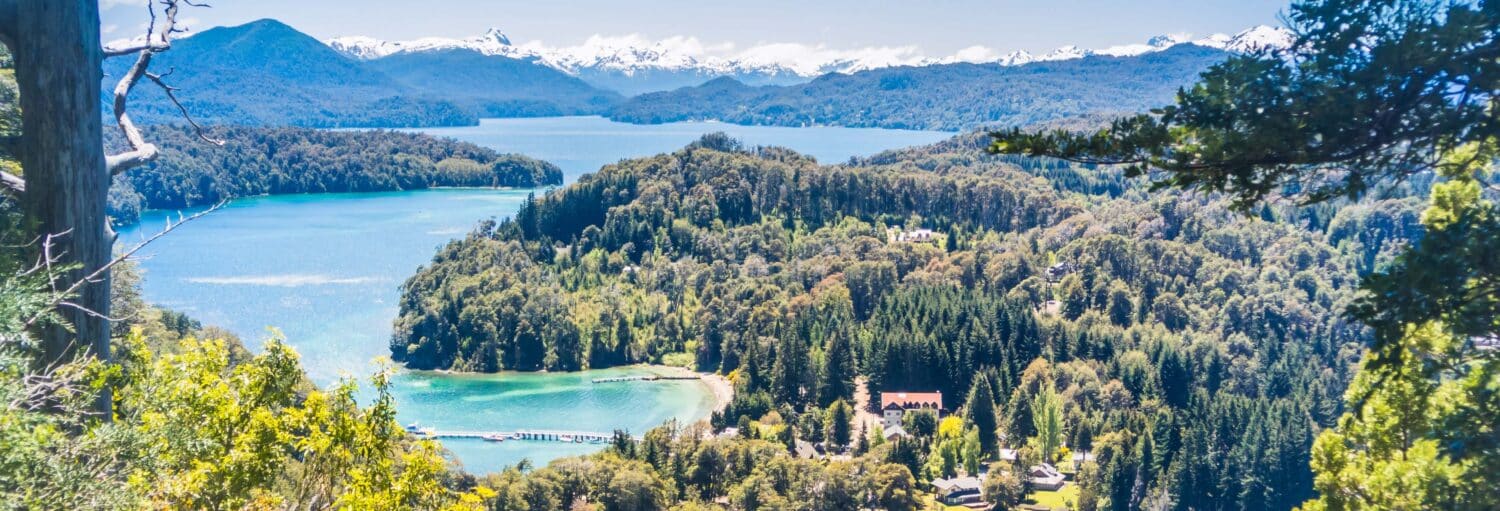 Uma península arborizada se estende em um lago azul com montanhas ao fundo sob um céu claro no Tour de neve pela Villa La Angostura e Cerro Bayo