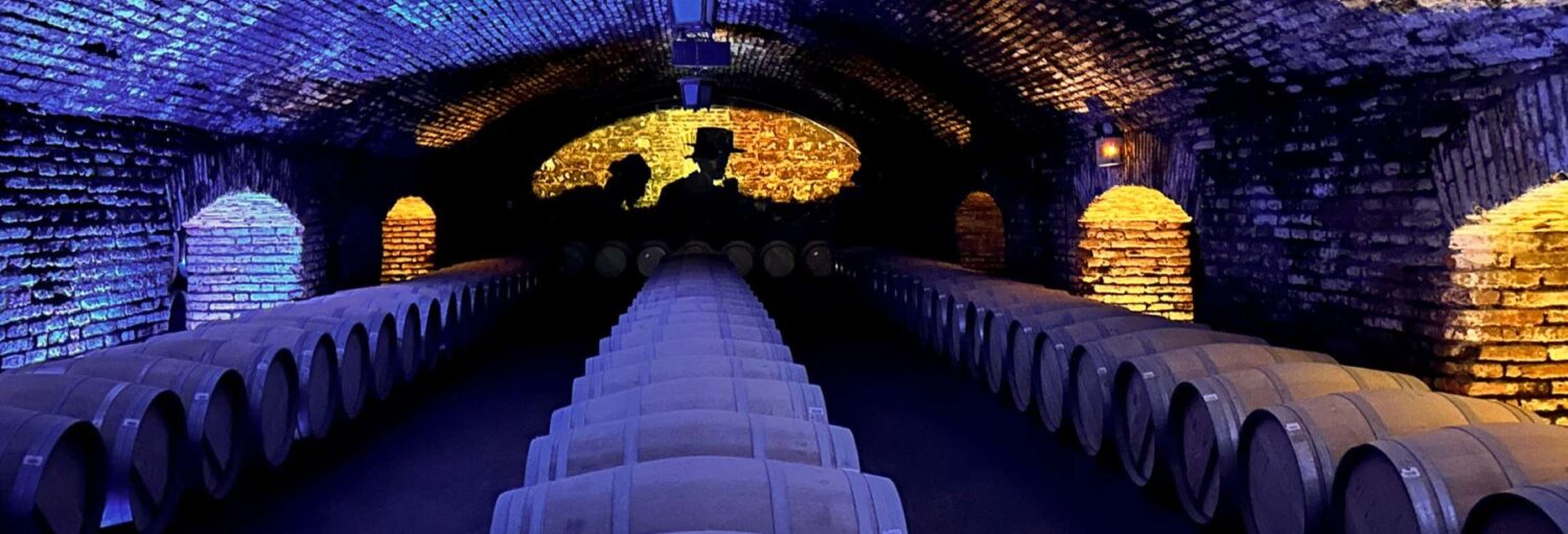 Uma adega pouco iluminada com fileiras de barris e paredes de tijolos iluminadas por luzes azuis e amarelas. A foto é capa do post do Tour do vinho Casillero del Diablo na Vinícola Concha y Toro + Jantar.