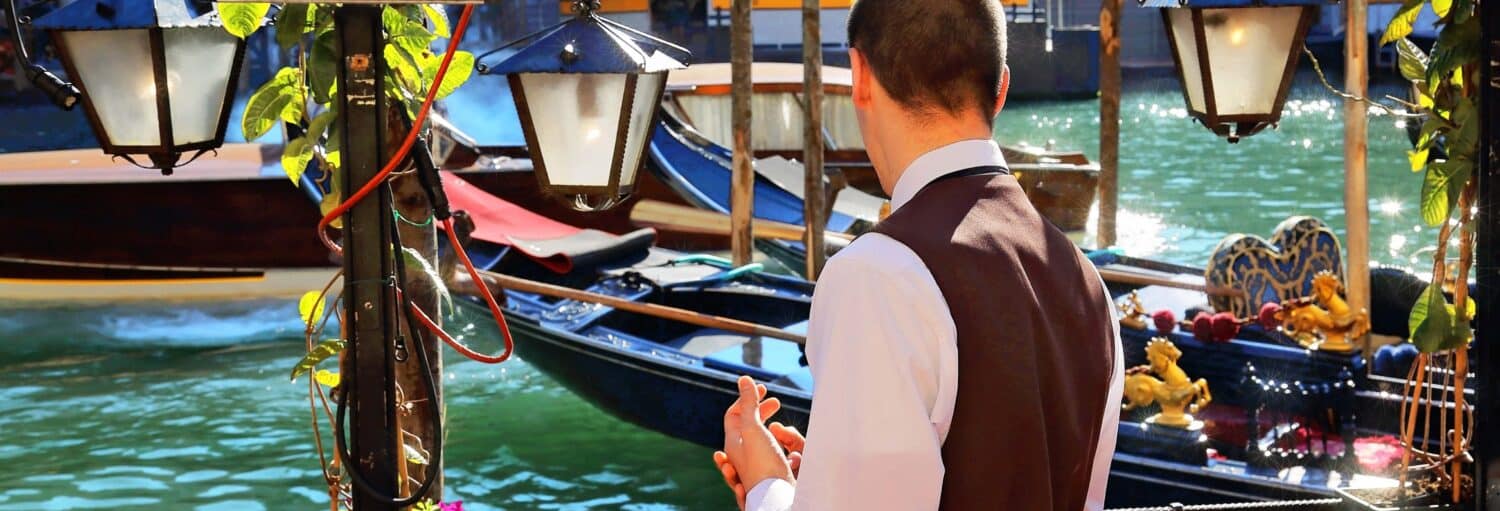 Um homem de colete desfruta de uma degustação de vinhos venezianos pendurando lâmpadas perto de barcos no canal em um dia ensolarado.