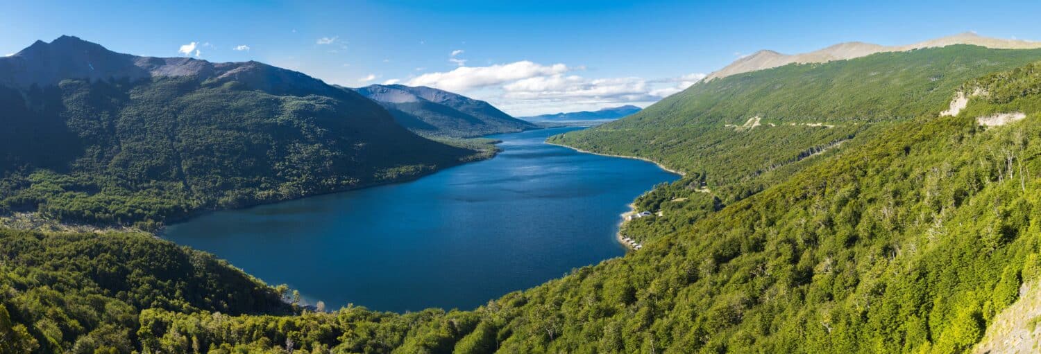 Um rio largo se curva entre montanhas verdes sob um céu azul com nuvens dispersas. Imagem para ilustrar post sobre o tour do whisky pela Terra do Fogo.