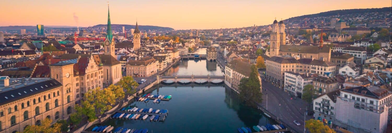 Vista aérea de Zurique com edifícios históricos, torres de igrejas e barcos ancorados no rio ao pôr do sol. Imagem para ilustrar o post sobre tour por Zurique ao entardecer + jantar de fondue.