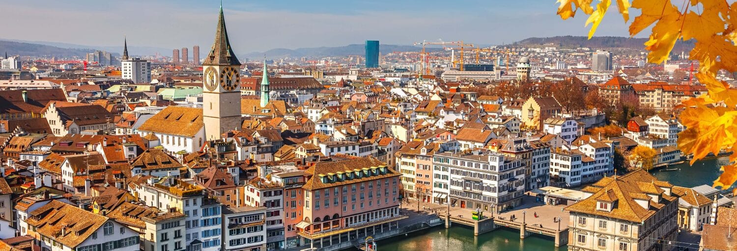Vista panorâmica do centro histórico da cidade de Zurique com a torre da igreja, o rio e as folhas de outono em primeiro plano. Imagem para ilustrar post sobre o tour por Zurique + passeio de barco + ingresso do Museu Lindt.