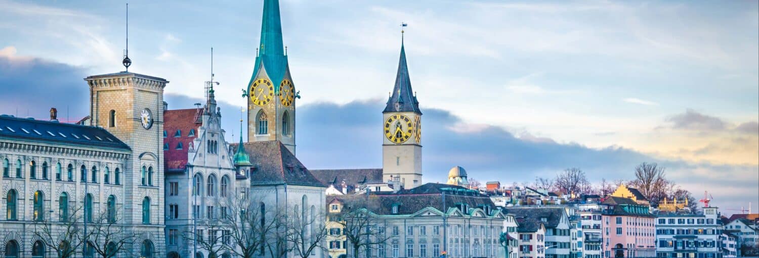 Paisagem urbana com edifícios históricos e torres de relógio sob um céu nublado em Zurique, Suíça. Imagem para ilustrar post sobre o tour por Zurique + ferry + teleférico de Felsenegg.