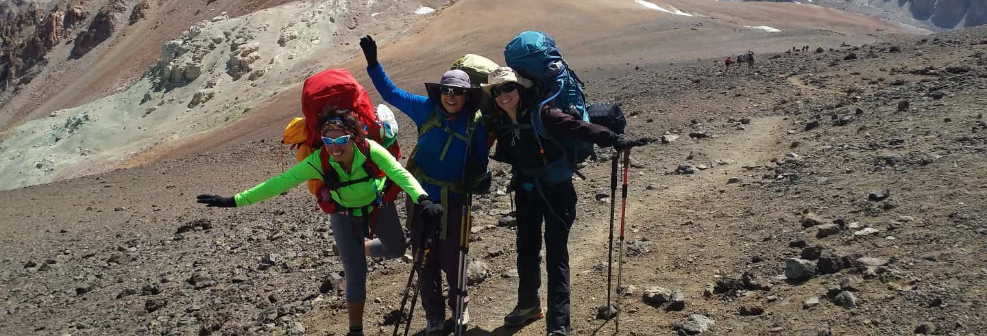 Três caminhantes com grandes mochilas e bastões de caminhada estão em uma trilha rochosa na montanha, sorrindo e posando para a câmera.