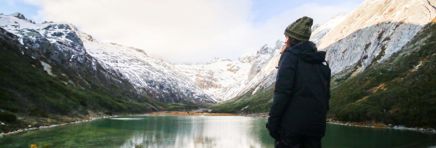 Uma pessoa vestida com roupas de inverno está ao lado de um lago de montanha cercado por picos cobertos de neve e colinas verdes. Imagem para ilustrar post sobre a trilha ao entardecer até a Laguna Esmeralda.