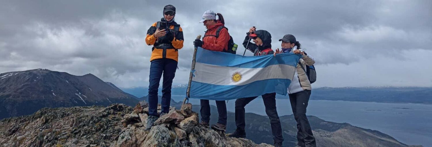 Quatro caminhantes estão em um pico de uma montanha rochosa, segurando uma bandeira argentina, com o céu nublado e um corpo de água distante atrás deles. Imagem para ilustrar post sobre trilha pelo Cerro Cortez.