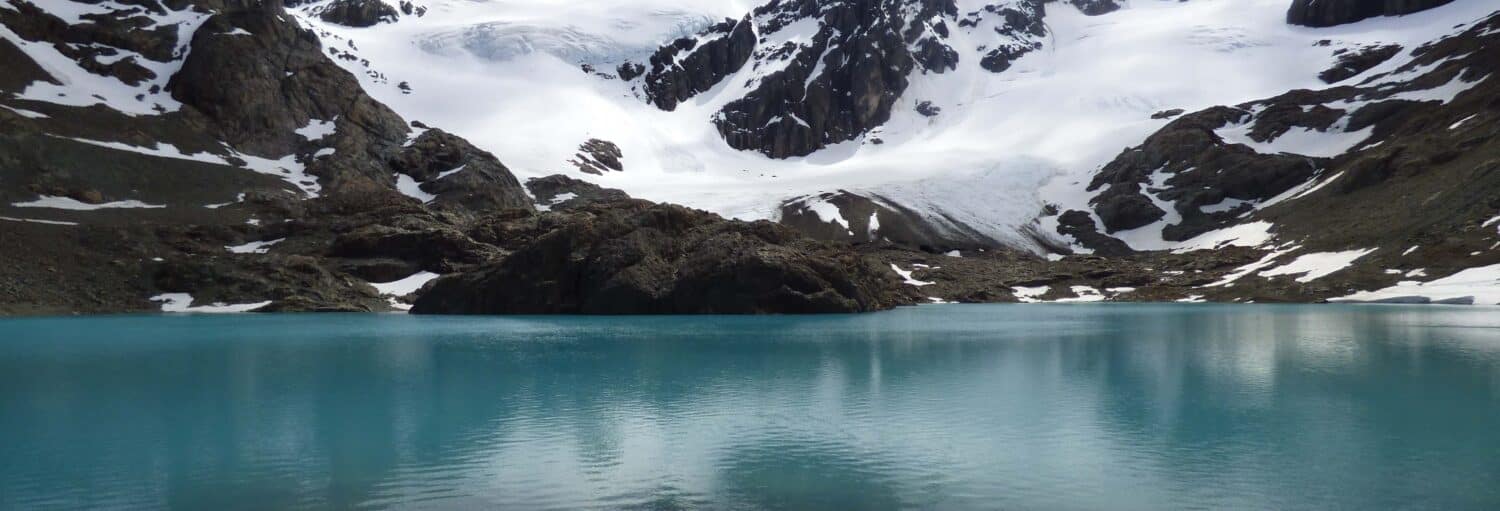 Um lago glacial azul-turquesa é cercado por montanhas rochosas cobertas de neve sob um céu nublado. Imagem para ilustrar um post sobre trilha pelo glaciar Vinciguerra.