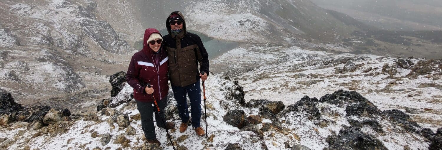 Duas pessoas com jaquetas de inverno e óculos escuros em pé em uma encosta de montanha nevada com terreno rochoso e fundo enevoado. Imagem para ilustrar post sobre a trilha pela Laguna Turquesa e pelo Monte Carbajal.