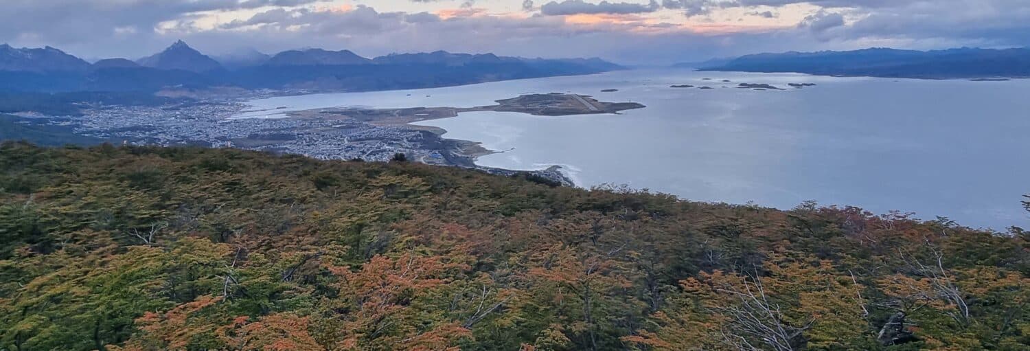 Uma cidade litorânea com uma baía, cercada por montanhas e floresta densa sob um céu nublado ao pôr do sol. Imagem para ilustrar post sobre a trilha ao entardecer pelo Monte Susana.