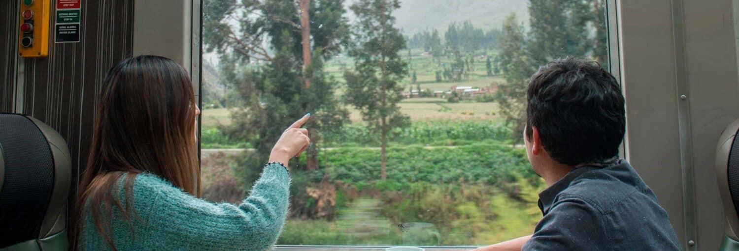 Duas pessoas sentadas em um trem, olhando pela janela para campos verdes e árvores; uma delas está apontando para fora no Trem a Cusco