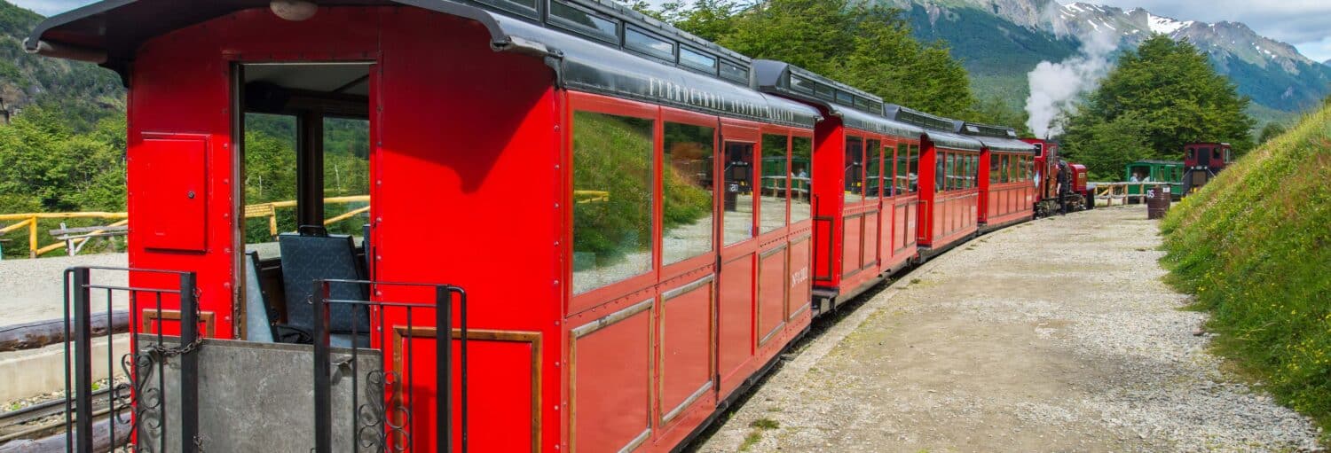 Um trem de passageiros vermelho brilhante em um trilho curvo com árvores verdes e montanhas ao fundo. Imagem para ilustrar post sobre o Trem do Fim do Mundo com audioguia.