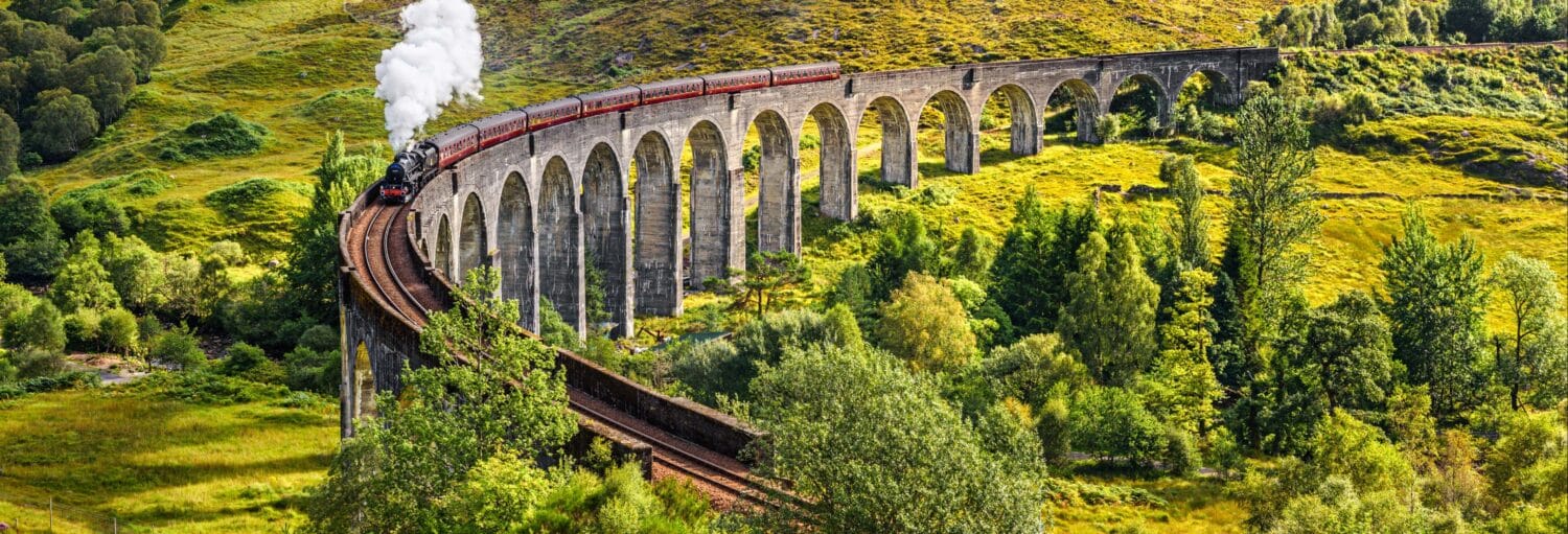 Um trem a vapor atravessa um longo viaduto de pedra cercado por colinas verdes e árvores em um dia ensolarado. A foto é capa do post do Trem do Harry Potter.