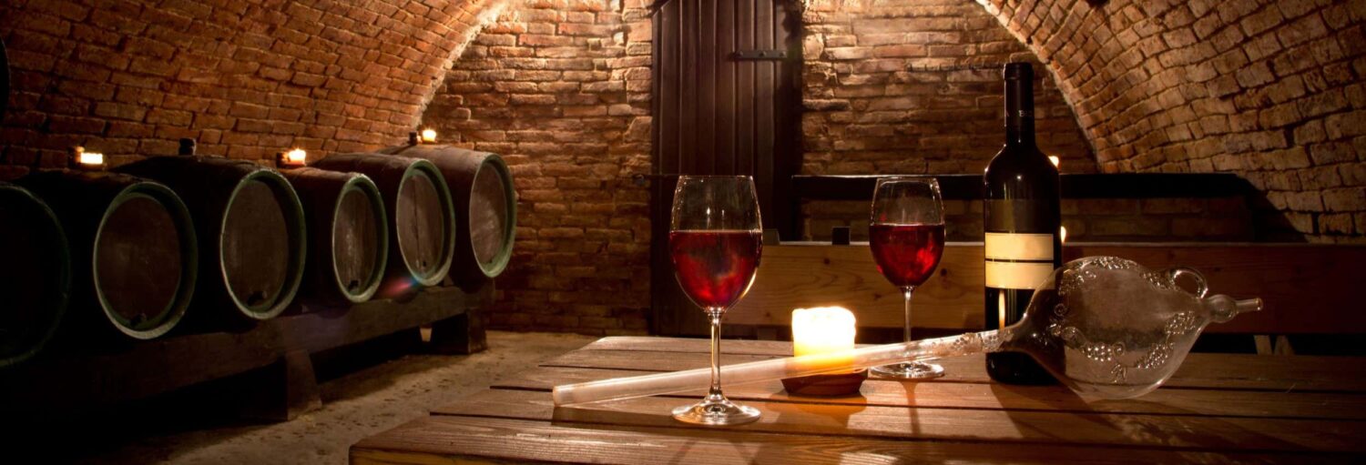 Uma adega mal iluminada com barris, duas taças de vinho tinto, uma garrafa, uma vela e um decantador de vidro em uma mesa de madeira. Imagem para ilustrar post sobre visita à vinícola Juanicó.