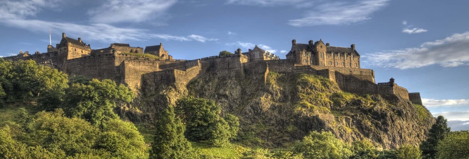 Um grande castelo de pedra fica no topo de uma colina rochosa, cercado por árvores verdes sob um céu azul com nuvens dispersas. A foto é capa do post da Visita guiada pelo Castelo de Edimburgo.