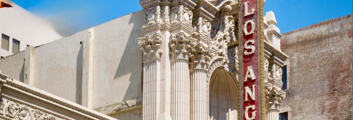 Fachada de teatro ornamentada com colunas altas e um letreiro vertical vermelho com a inscrição "Los Angeles" contra um céu azul. A foto é capa do post da Visita guiada pelo Downtown de Los Angeles.