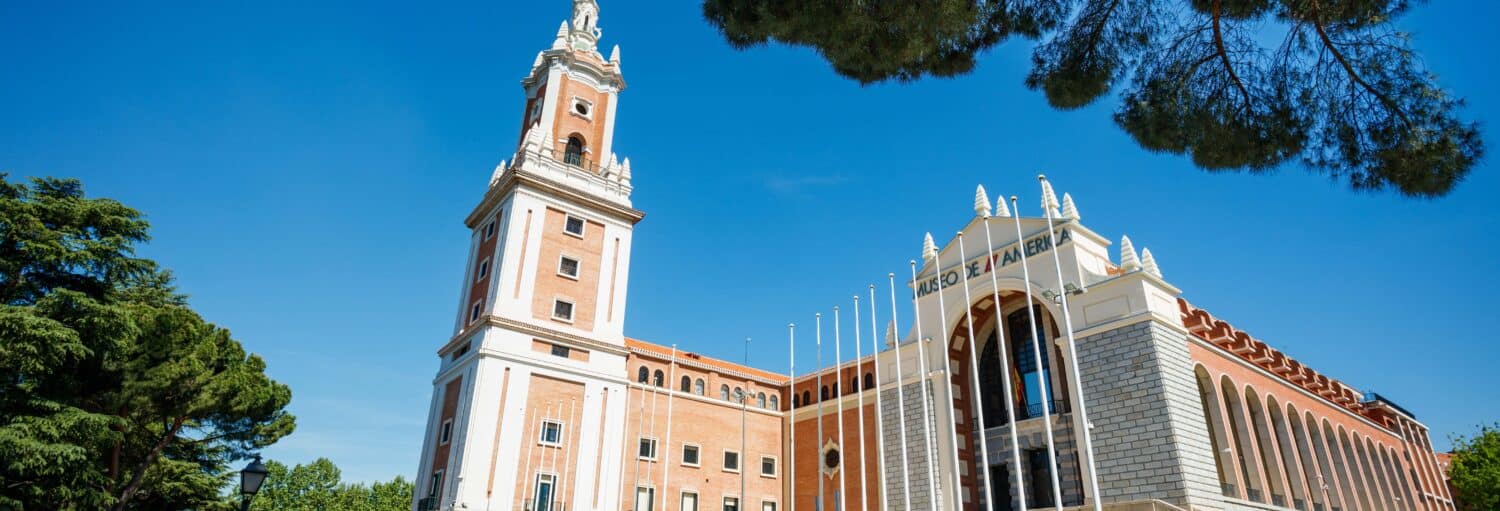 Um grande edifício de tijolos e pedras com uma alta torre de relógio, janelas em arco e vários mastros de bandeira sob um céu azul. A imagem ilustra o post sobre Visita guiada pelo Museu da América.