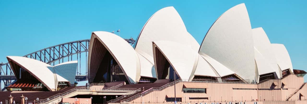 Sydney Opera House com seus telhados brancos em forma de vela, com a Sydney Harbour Bridge visível ao fundo em um céu claro. Imagem para ilustrar post sobre a visita guiada pela Ópera de Sydney.
