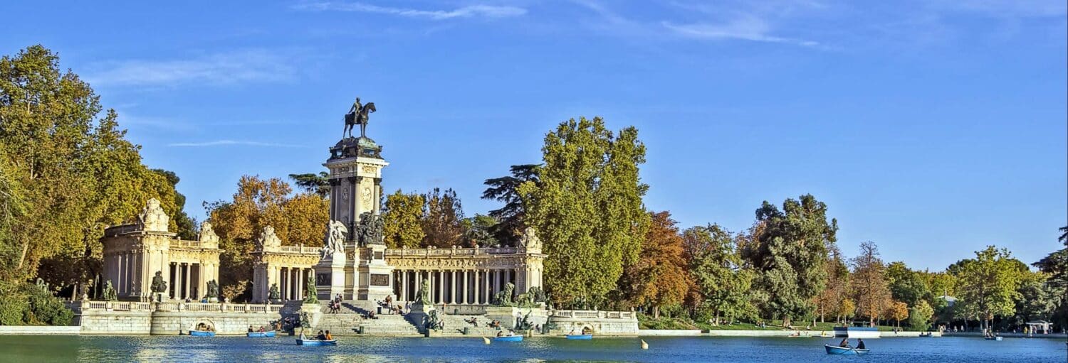 Um grande monumento com uma estátua de cavalo tem vista para um lago com pessoas passeando de barco, cercado por árvores sob um céu azul. a imagem ilustra o post sobre Visita guiada pelo Parque El Retiro.