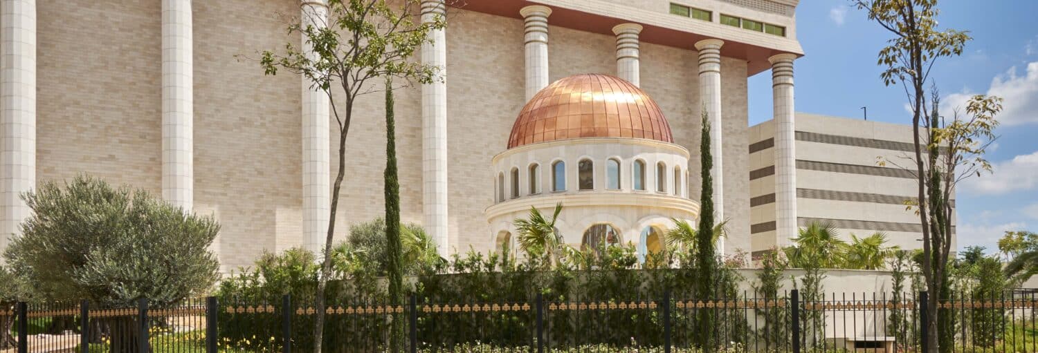 Um edifício branco abobadado com colunas e palmeiras, cercado por uma cerca e um jardim sob um céu azul. a imagem ilustra o post sobre Visita guiada pelo Templo de Salomão.