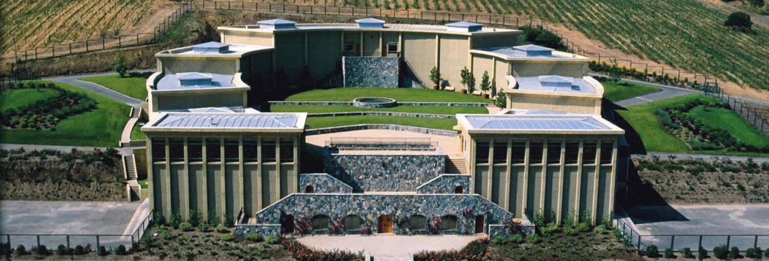 Um edifício grande e moderno com detalhes em pedra e jardins paisagísticos, situado próximo a campos em um declive suave. A foto é capa do post da Visita à vinícola Haras de Pirque.