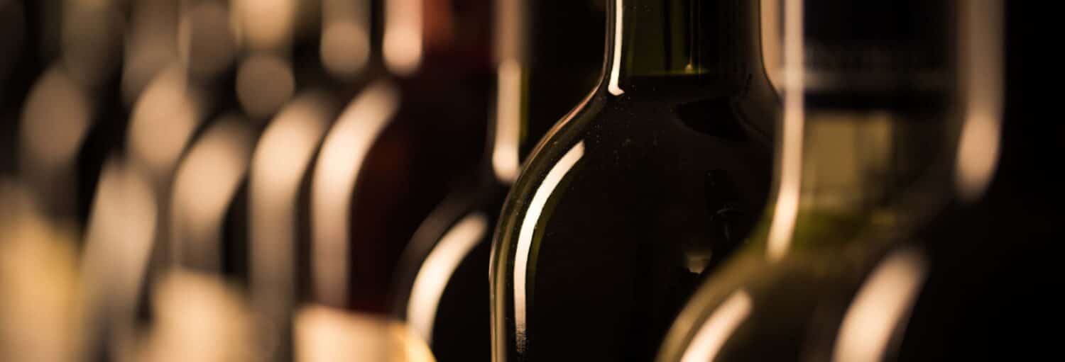 Close-up de várias garrafas de vinho alinhadas em uma fileira com fundo desfocado e iluminação quente da Visita à vinoteca Wine House