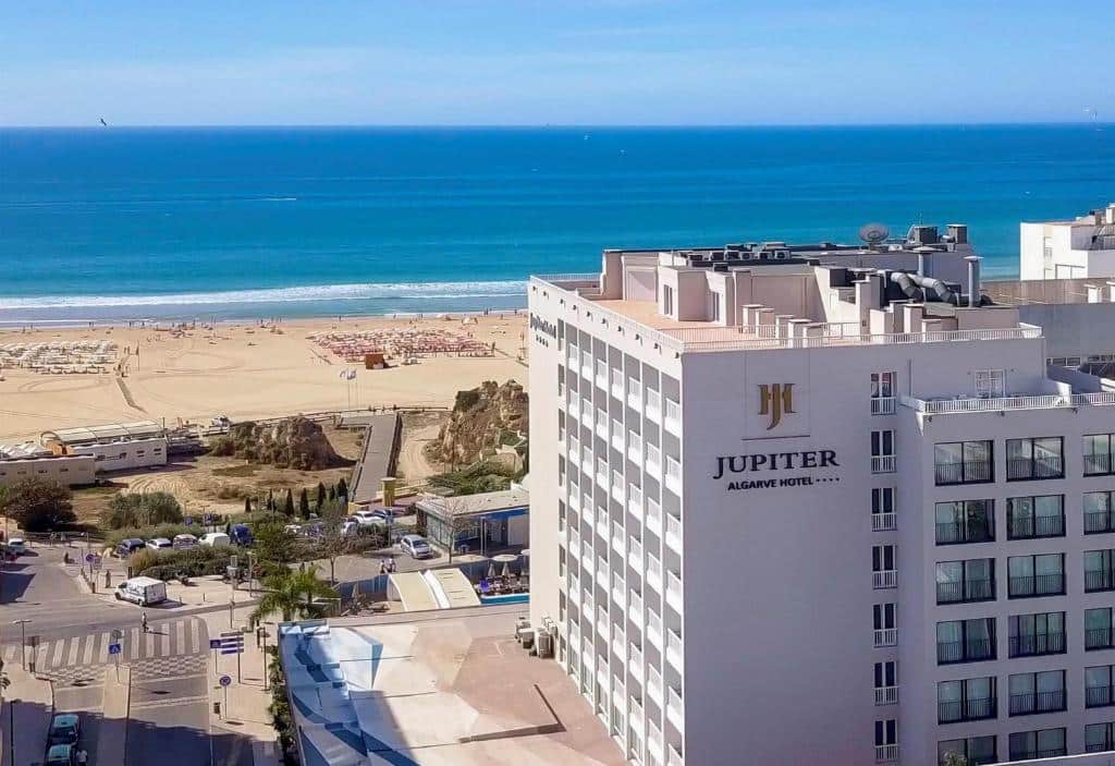Um grande hotel chamado "Jupiter Algarve Hotel" fica perto de uma praia de areia e do oceano azul sob um céu claro.