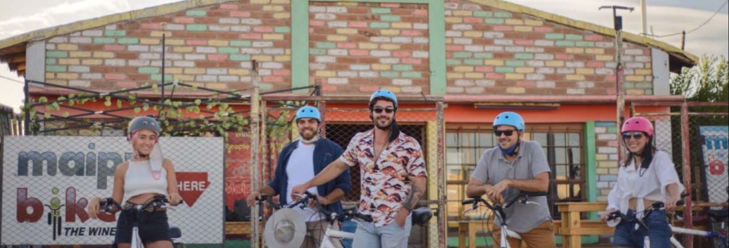 Cinco pessoas com capacetes estão paradas com bicicletas em frente a um prédio de tijolos coloridos e uma placa que diz "bicicleta principal" durante o Aluguel de bicicleta em Maipú