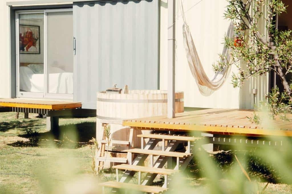 Um pátio da Las Guaras com uma banheira de hidromassagem de madeira, rede e degraus leva a um edifício moderno com grandes portas de vidro e um quarto visível.