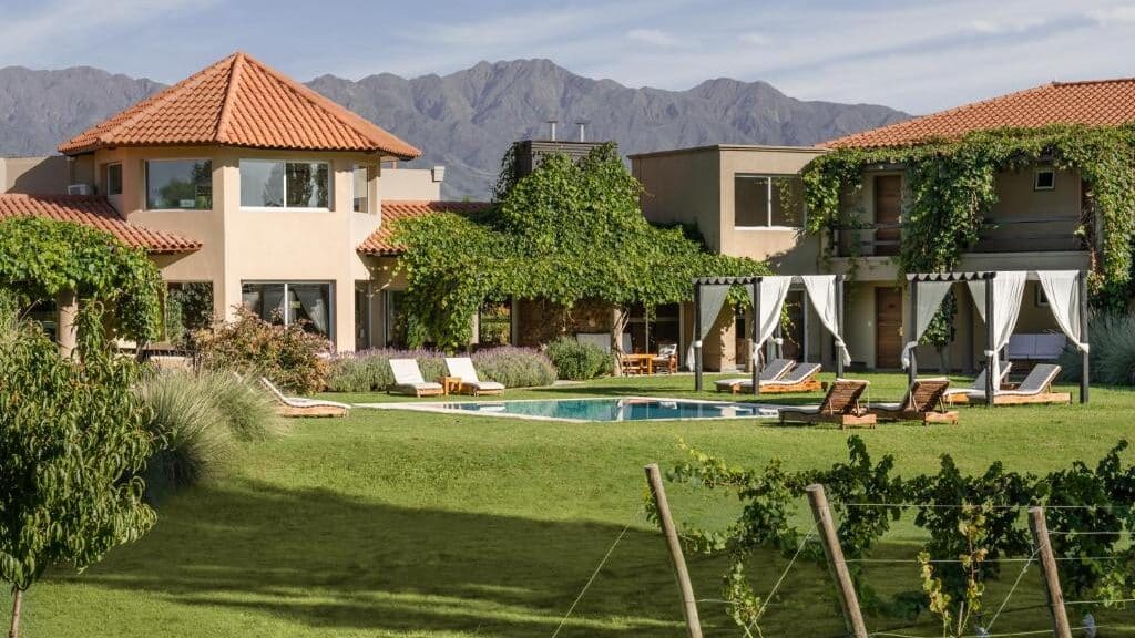 Villa Mansa com telhados de terracota, uma piscina, espreguiçadeiras e montanhas ao fundo, cercada por vegetação e videiras. Imagem para ilustrar post sobre onde ficar em Mendoza.