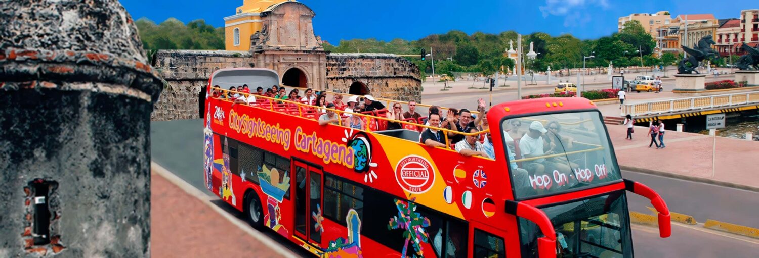 Um ônibus turístico vermelho de dois andares com turistas passa por muros e edifícios históricos de pedra sob um céu azul claro. Imagem para ilustrar post sobre o ônibus turístico de Cartagena.