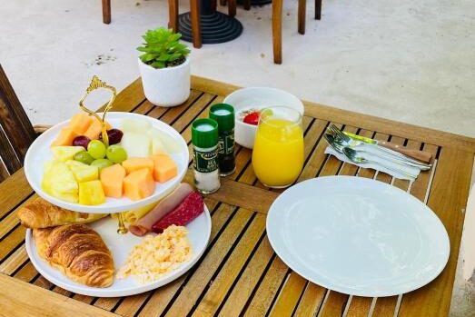Uma mesa de madeira do Atelier Skalo Boutique Hotel com um prato de frutas, croissant, ovos, suco, condimentos, garfos, facas e um prato vazio. Imagem para ilustrar post sobre onde ficar em Curaçao.