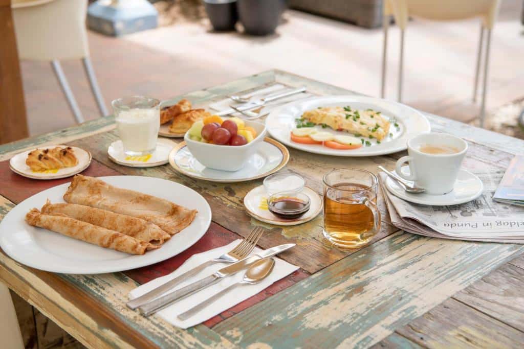 Uma mesa rústica do BOHO Bohemian Boutique Hotel com crepes, frutas, uma omelete, café, chá, suco, uma massa, utensílios e um jornal.