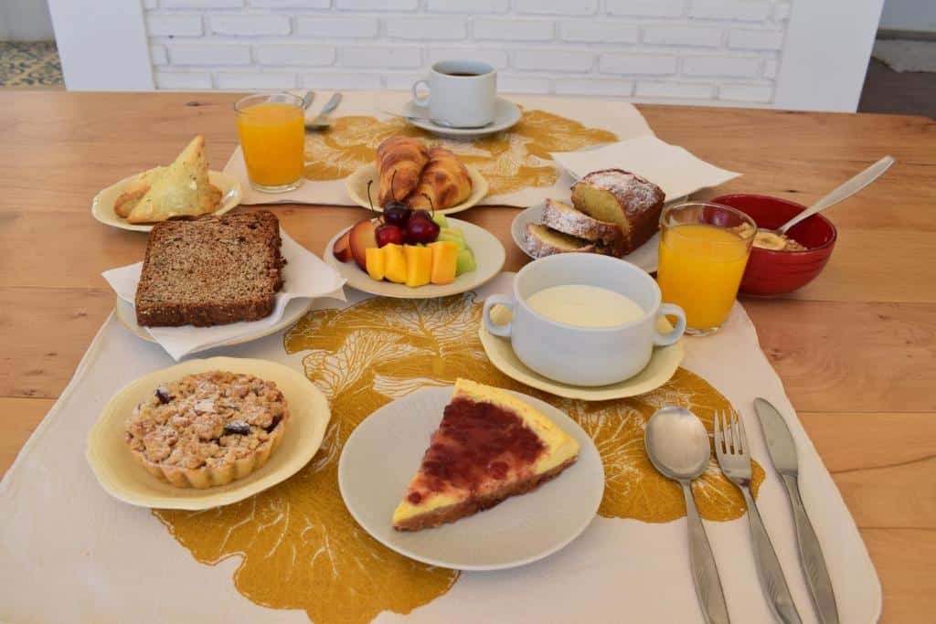 Café da manhã da Villa Mendotza com pães, bolos, frutas, suco de laranja, café e iogurte em uma mesa com um jogo de mesa de folhas amarelas.
