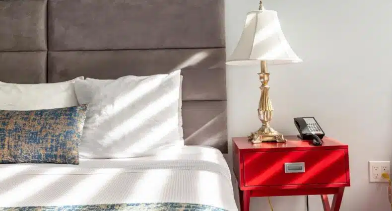 Uma cama com lençóis brancos e um travesseiro com estampa azul ao lado de uma mesa de cabeceira vermelha com um abajur e um telefone.