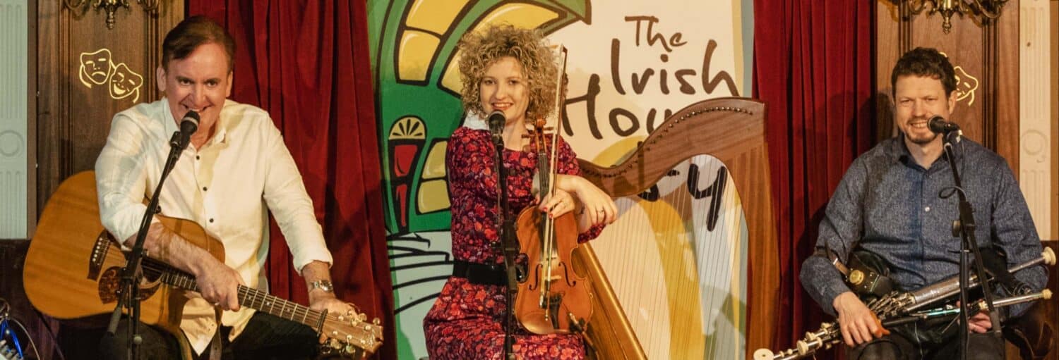 Três músicos no palco com violão, violino, harpa e gaitas, sentados em frente a uma placa que diz "The Irish Ho". A foto é capa do post do Jantar e espetáculo folclórico em Dublin.