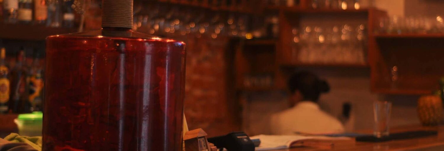 Um grande jarro de vidro vermelho fica em um balcão de bar com prateleiras de copos e uma pessoa ao fundo. Imagem para ilustrar post sobre o jantar em um restaurante do centro histórico.