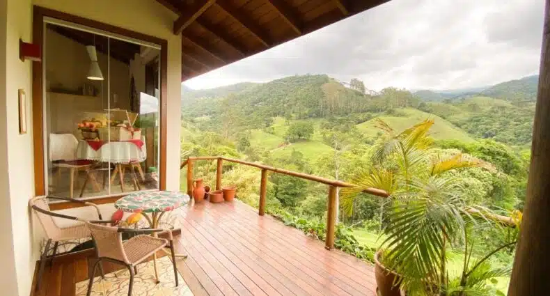 Varanda de madeira com duas cadeiras e uma mesa redonda com vista para as colinas verdes; a área de jantar interna é visível através de uma janela, na propriedade do Chalé nas montanhas em São Francisco Xavier, para representar chalés em São Francisco Xavier.