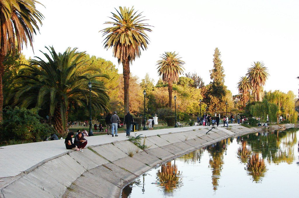 A imagem mostra um parque urbano à beira de um lago.O cenário é ao ar livre, com a luz do sol criando reflexos dourados na água. À direita, há um canal de concreto com água, que reflete as árvores e palmeiras que ficam na margem oposta. À esquerda do canal, há um caminho largo e pavimentado onde várias pessoas caminham ou estão sentadas.