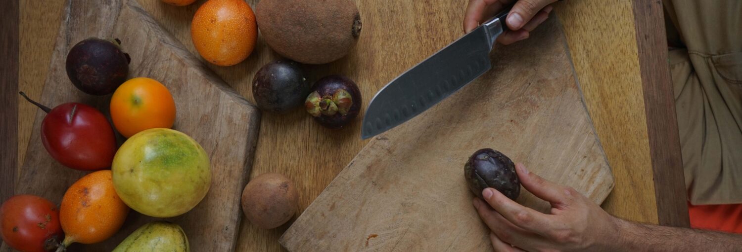 Uma pessoa corta uma fruta roxa escura em uma tábua de madeira, com várias frutas tropicais próximas em uma mesa. Imagem para ilustrar post sobre a degustação de maracujá em Cartagena.