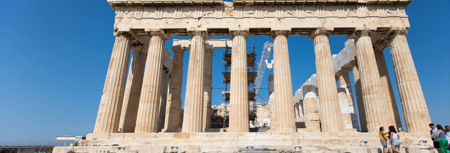 O Partenon, em Atenas, com os andaimes visíveis, fotografado em um dia claro com turistas à direita. A imagem ilustra o post sobre Ingresso da Acrópole de Atenas.