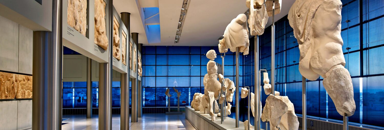 Estátuas e relevos de mármore branco são exibidos em uma moderna galeria de museu com paredes de vidro e iluminação azul. A imagem ilustra o post sobre Ingresso do Museu da Acrópole.