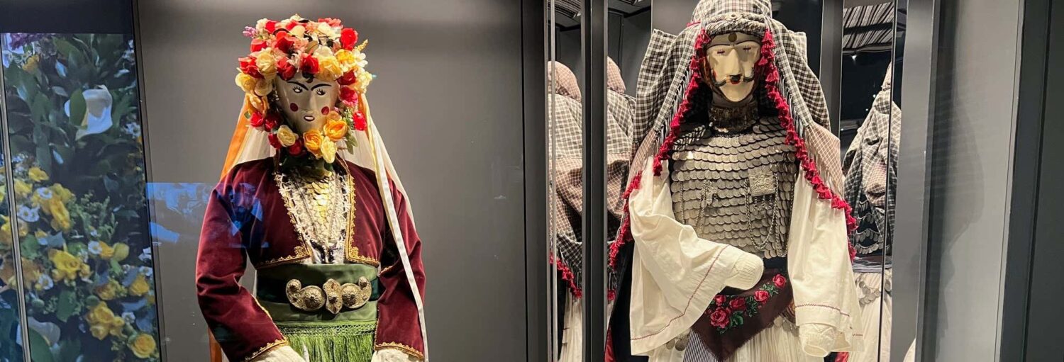 Dois manequins vestidos com trajes tradicionais com detalhes florais e de metal são exibidos em uma caixa de vidro. A imagem ilustra o post sobre Ingresso do Museu da Cultura Grega Moderna.