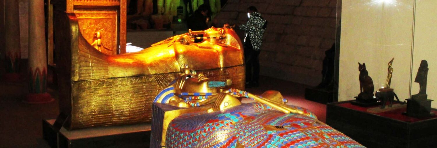 Um sarcófago e uma máscara de faraó de ouro exibidos em um museu com visitantes observando ao fundo. - Ingresso do Museu Egípcio de Canela