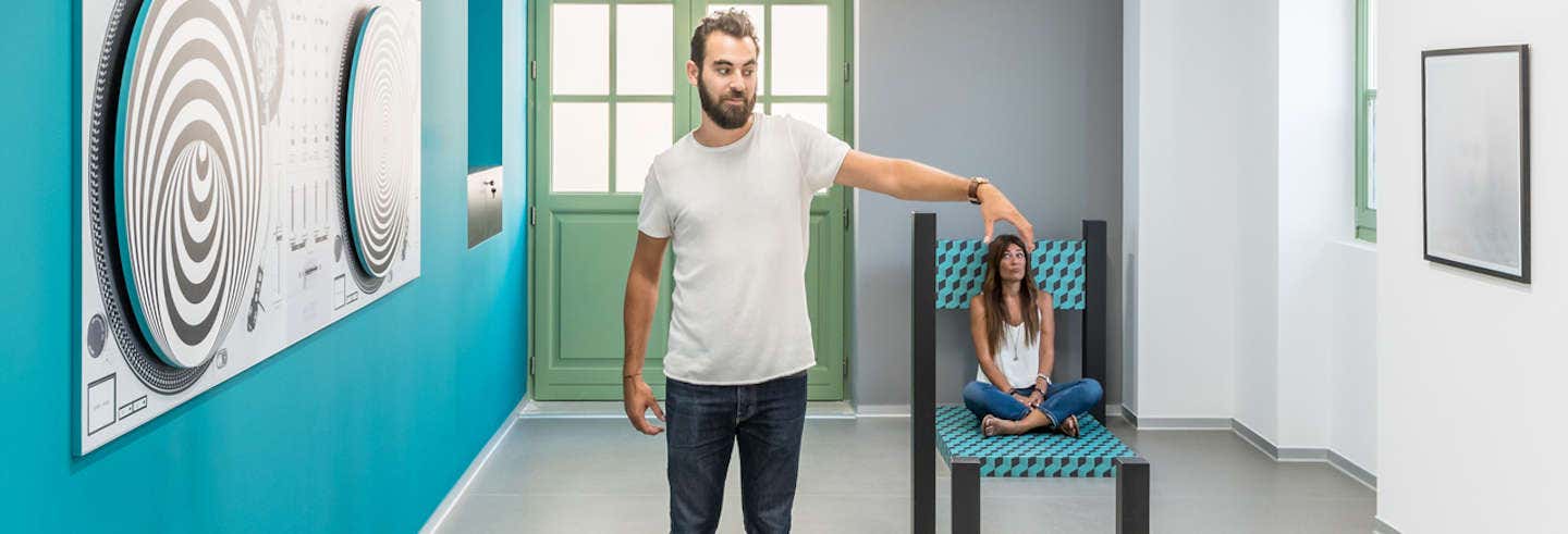 Um homem está de pé tocando o topo de uma cadeira enorme enquanto uma mulher se senta nela em uma sala bem iluminada com paredes azuis. A imagem ilustra o post sobre Ingresso do Museu das Ilusões de Atenas.